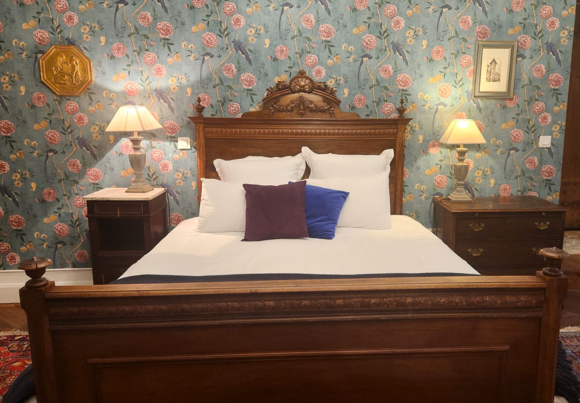 Chambre avec lit en bois orné, papier peint fleuri et lampes de chevet.