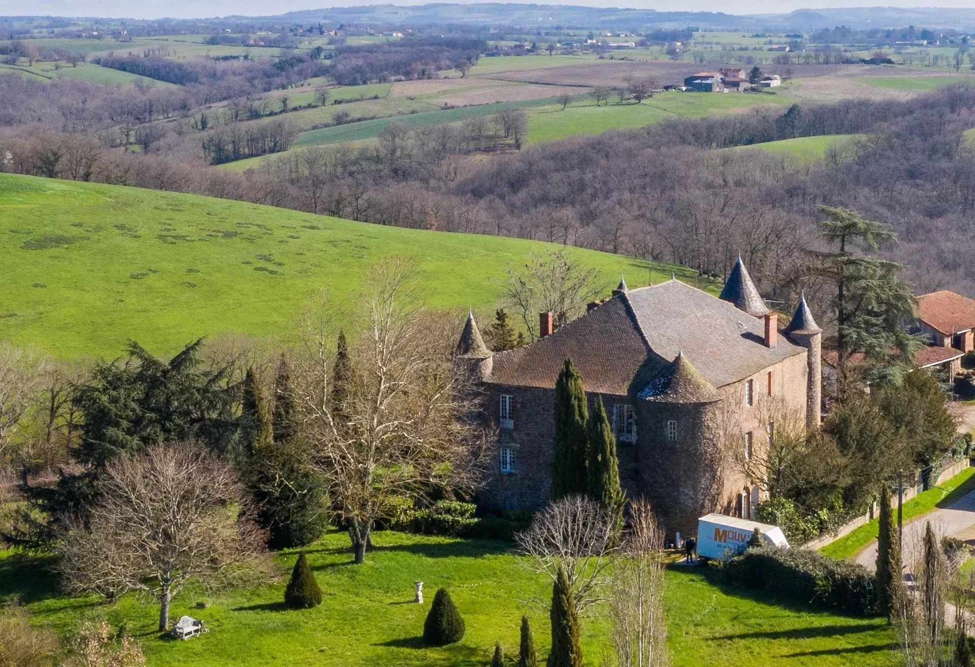 Château on a green lawn with a rural valley backdrop. Brown and green landscape with trees and a cloudy sky.