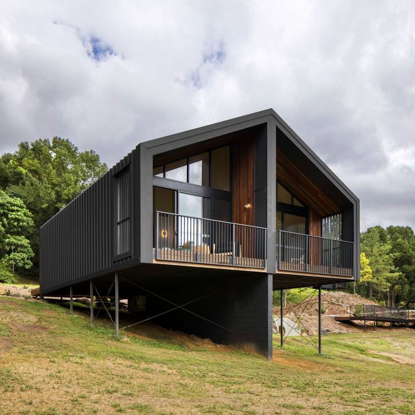 Modern black house with a balcony, built on stilts on a grassy hill, under a cloudy sky.
