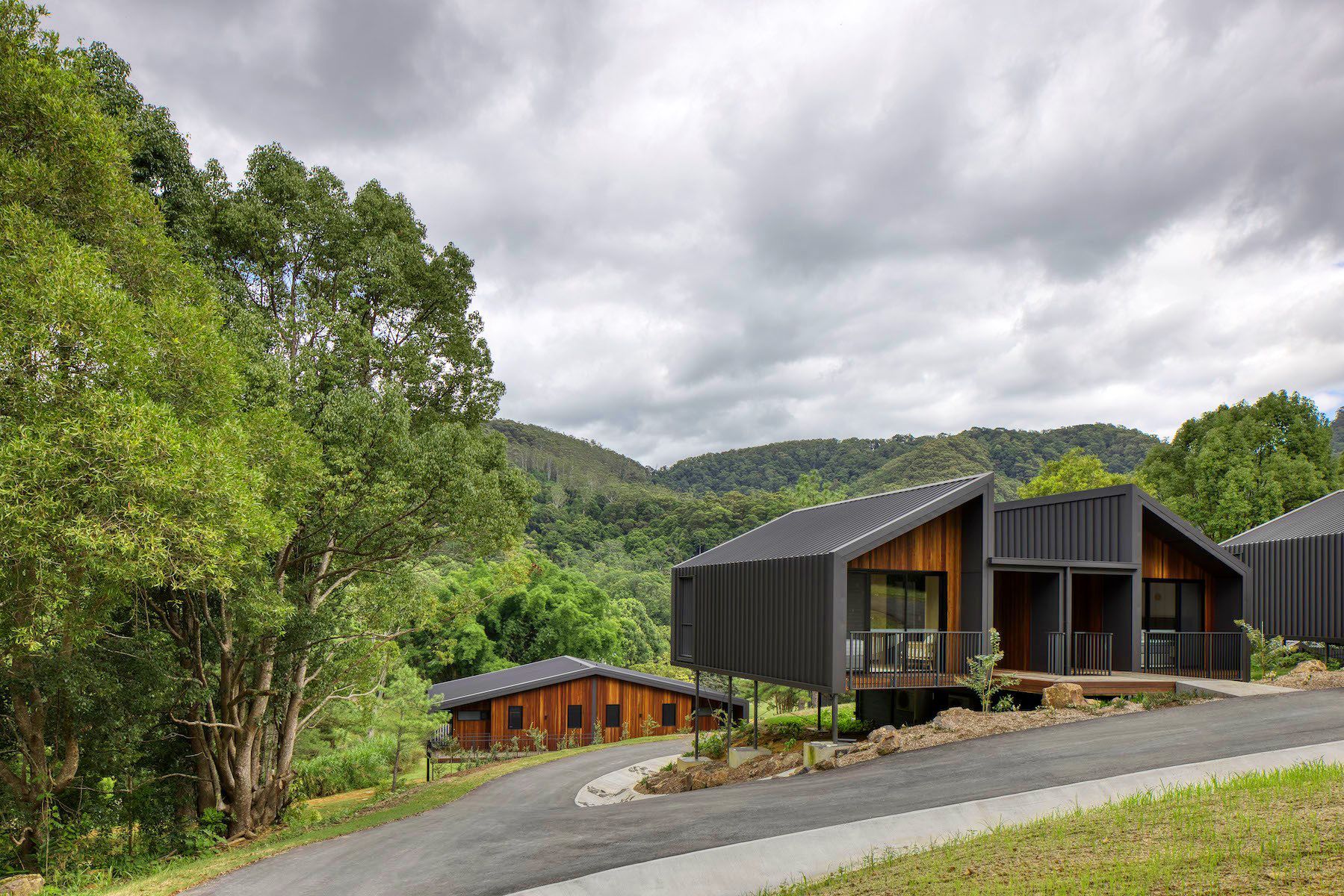 Modern cabins nestled on a hillside, with a winding road and lush green trees. Cloudy sky.
