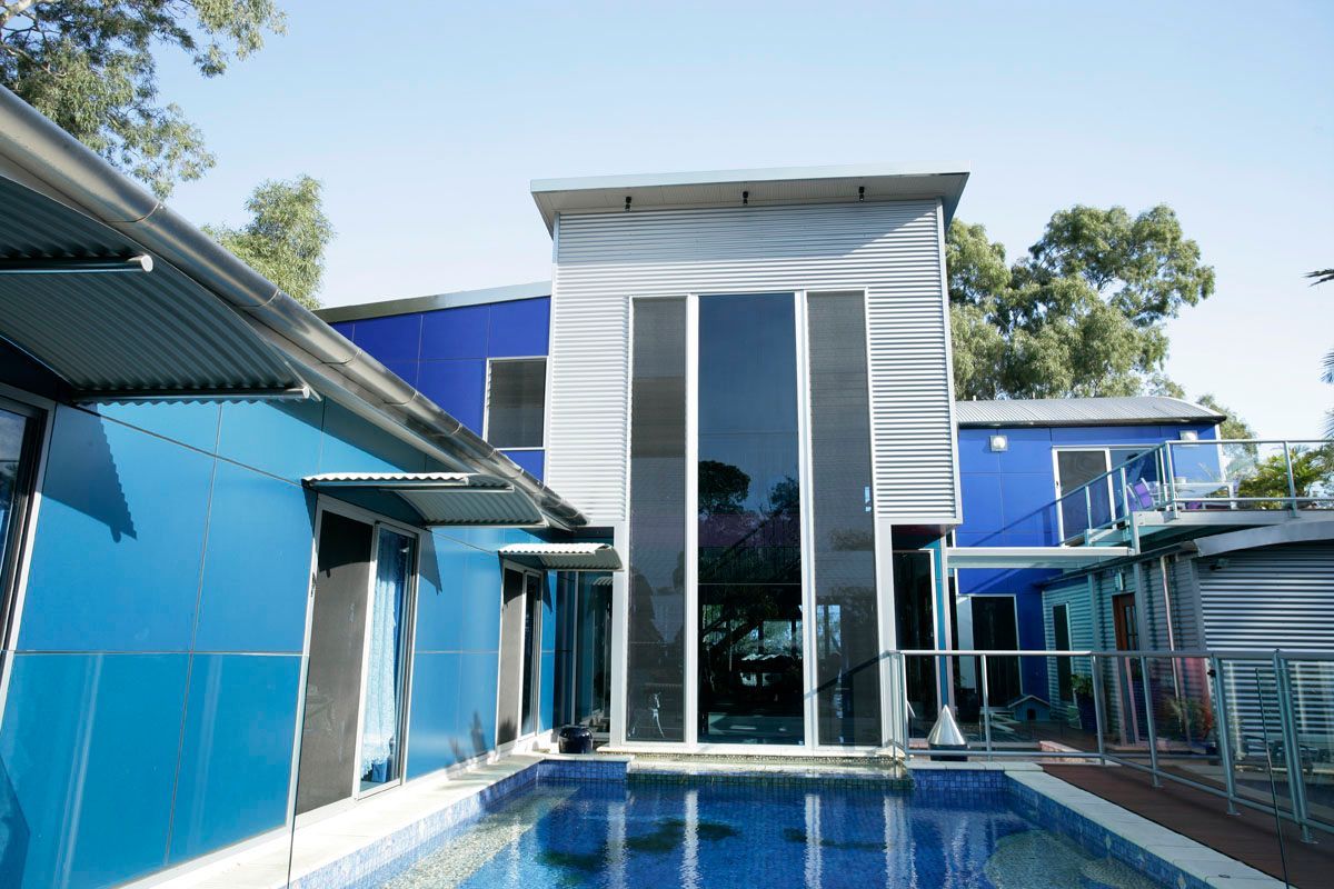 Blue and grey container home with pool. Tall windows, balcony, sunny outdoor setting.