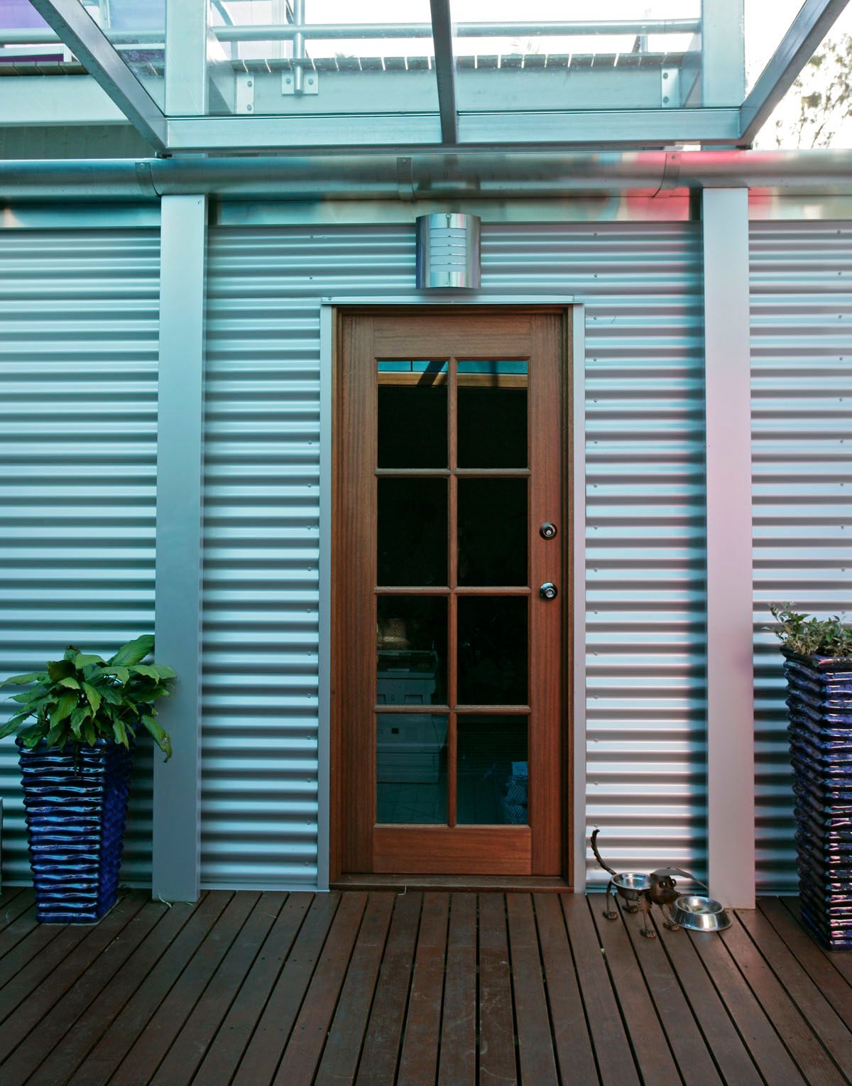 Wooden door with glass panes set in corrugated metal wall, on wooden deck with potted plants.