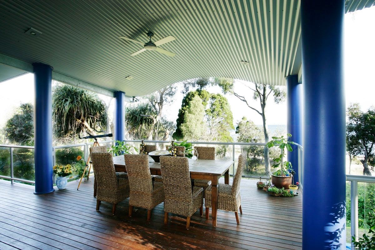 Outdoor dining area with blue columns, wooden deck, dining table, and chairs. Overlooks greenery and water.