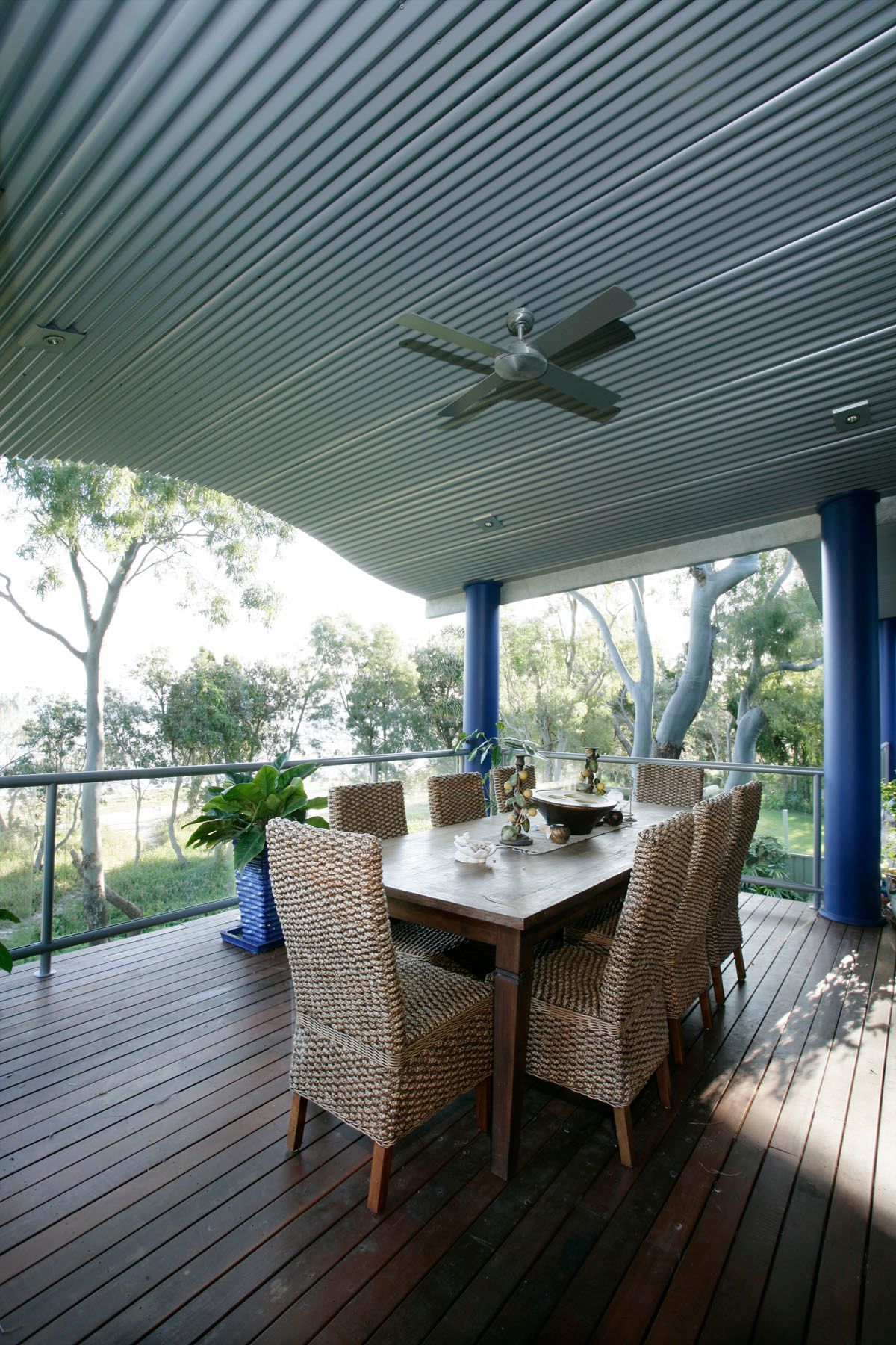 Outdoor dining area with wooden table, wicker chairs, and a blue-ceilinged patio overlooking a green landscape.
