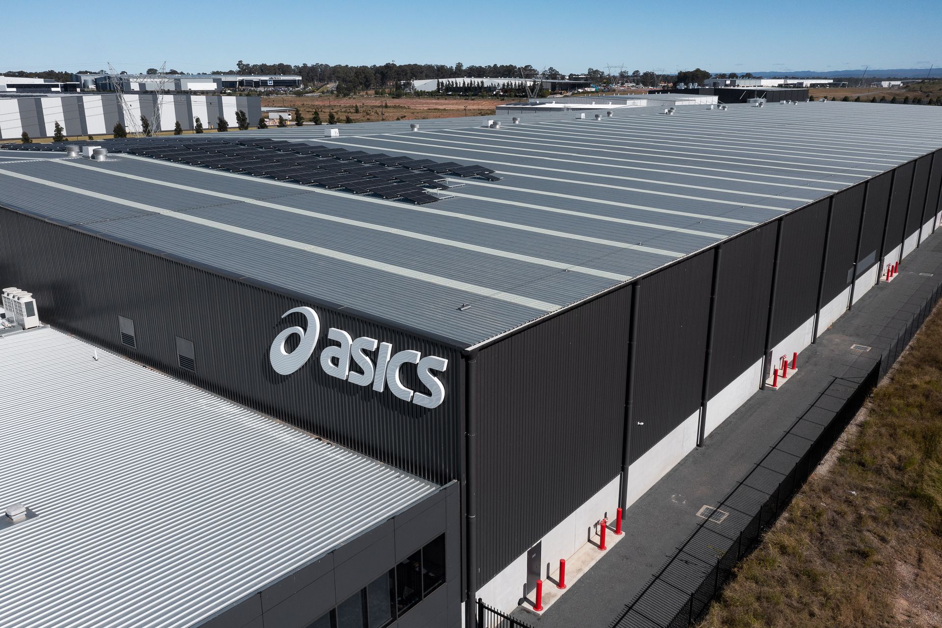 Large Asics warehouse with solar panels, gray and black exterior, daytime, logo visible.