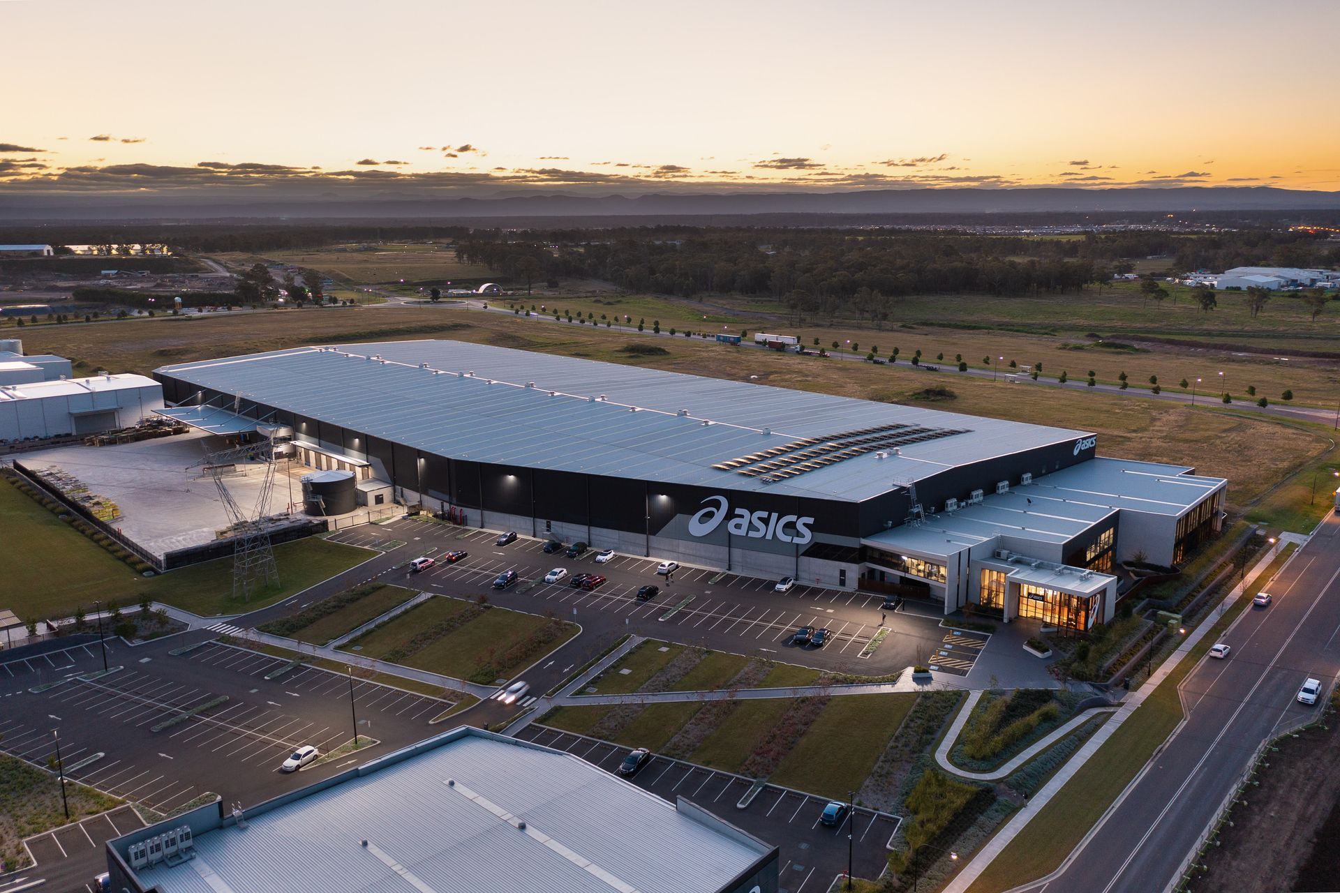 Aerial view of ASICS distribution center at sunset with large parking lot and highway.
