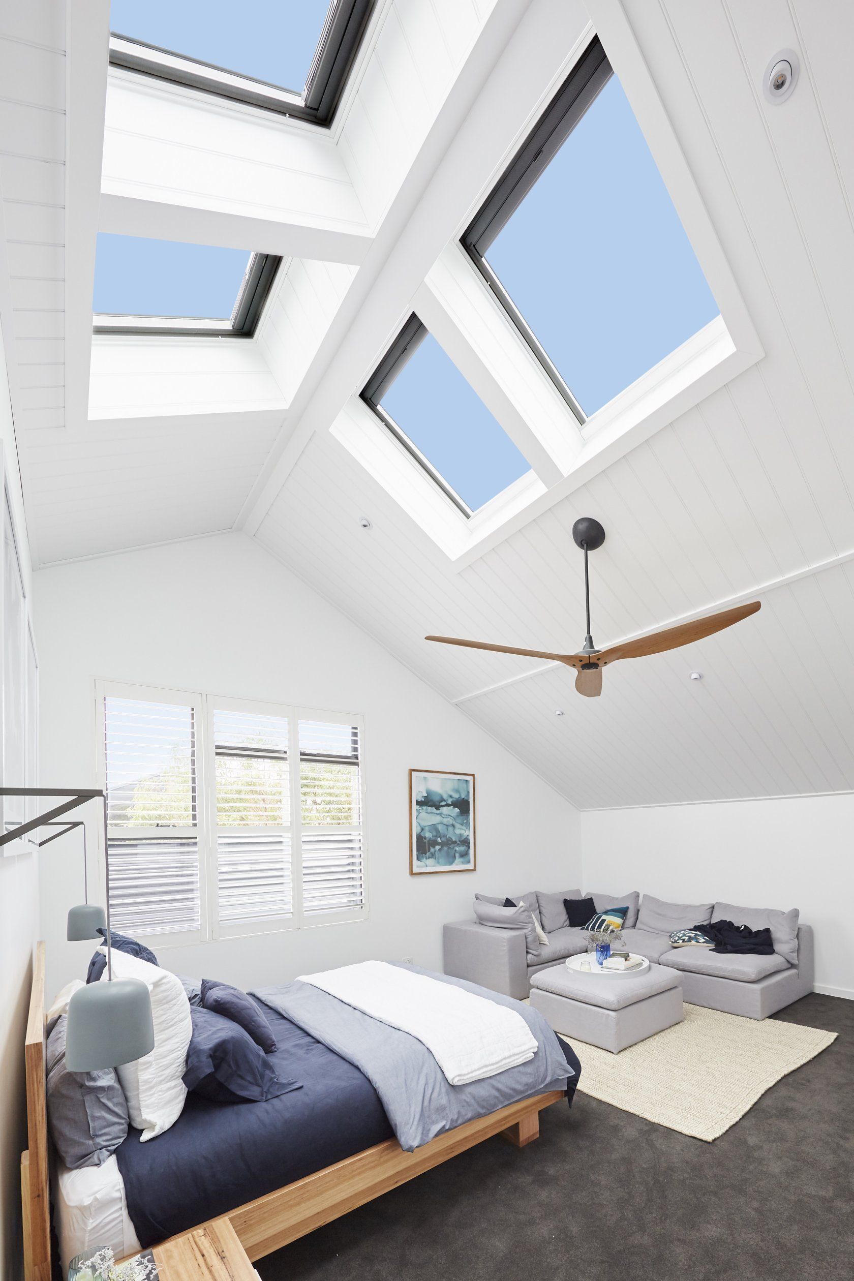 Bedroom with skylights, bed, sofa, and ceiling fan; white walls, blue sky visible through windows.