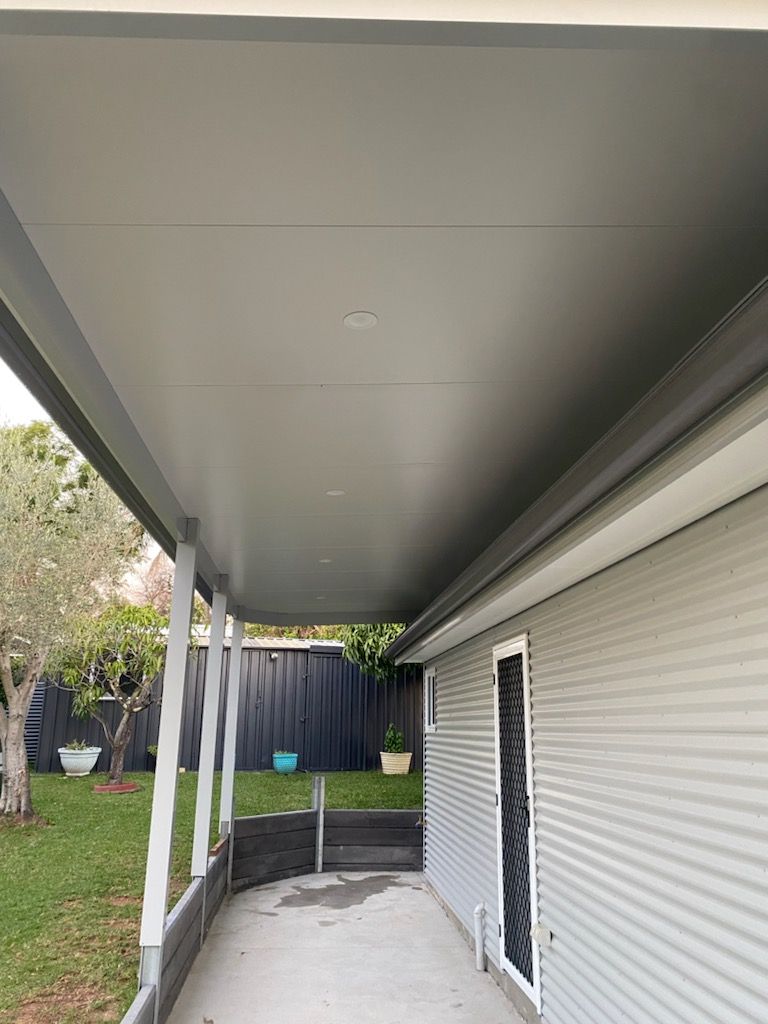 A covered patio with a gray roof, supported by white posts, next to a gray corrugated metal wall.