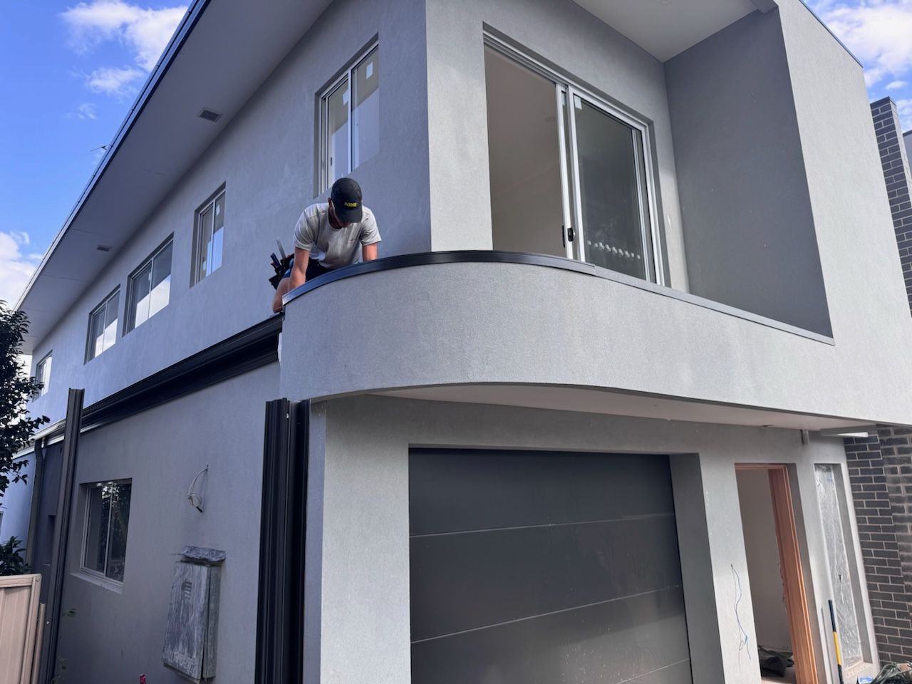 Man working on a modern two-story house's roof and balcony. The building is light grey with dark trim.