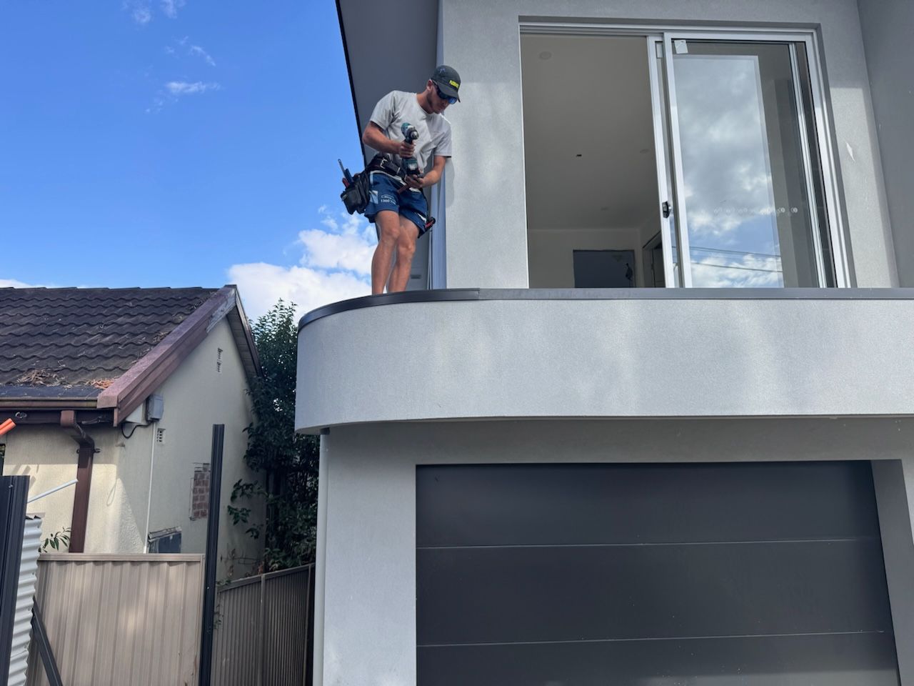 Man on roof, wearing a tool belt, working near an open balcony door. House with a gray garage door and blue sky.