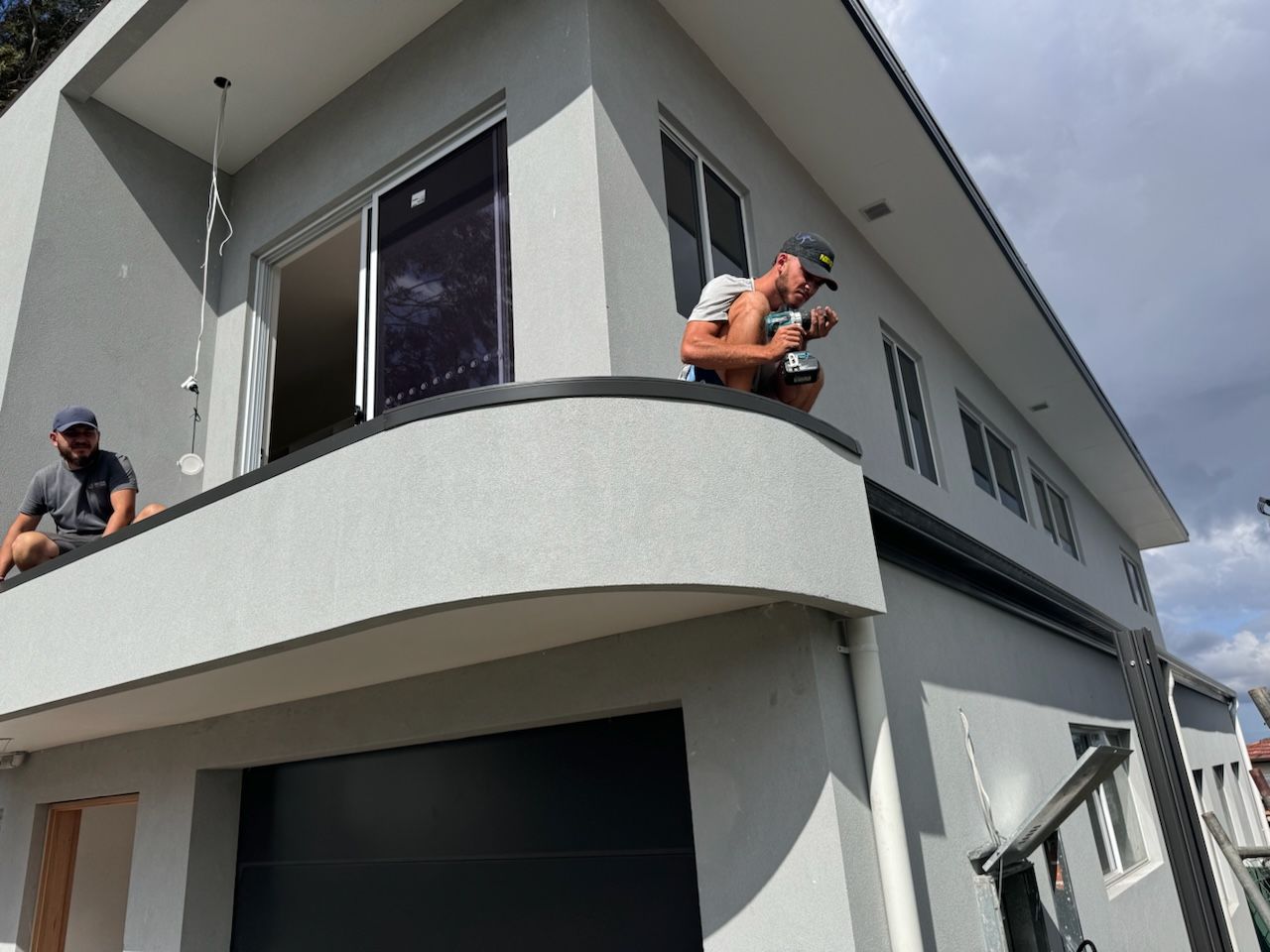 Two men working on a light gray house with black trim and garage door; one on a balcony.