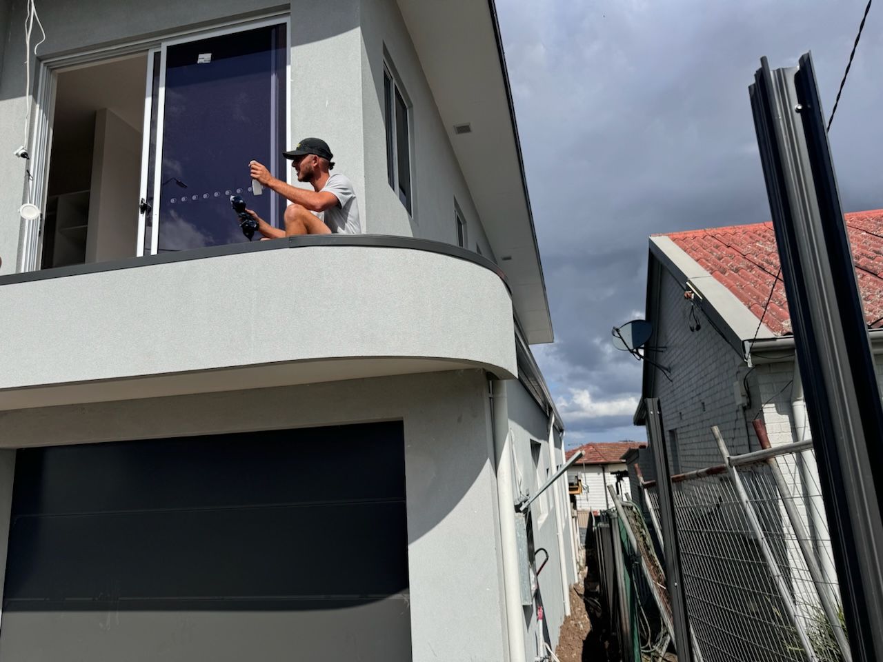 A man working on a balcony, spraying something. The house is gray and white. Overcast sky.