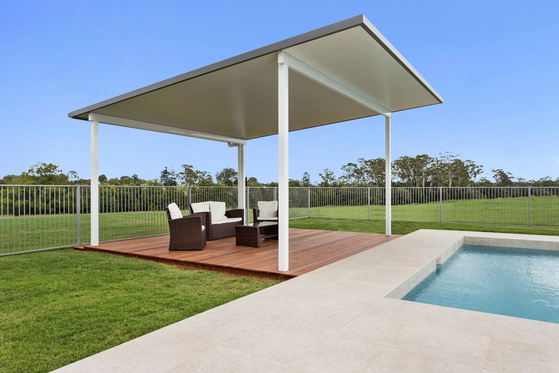 Patio with seating under a white-framed roof, near a pool on a sunny day.