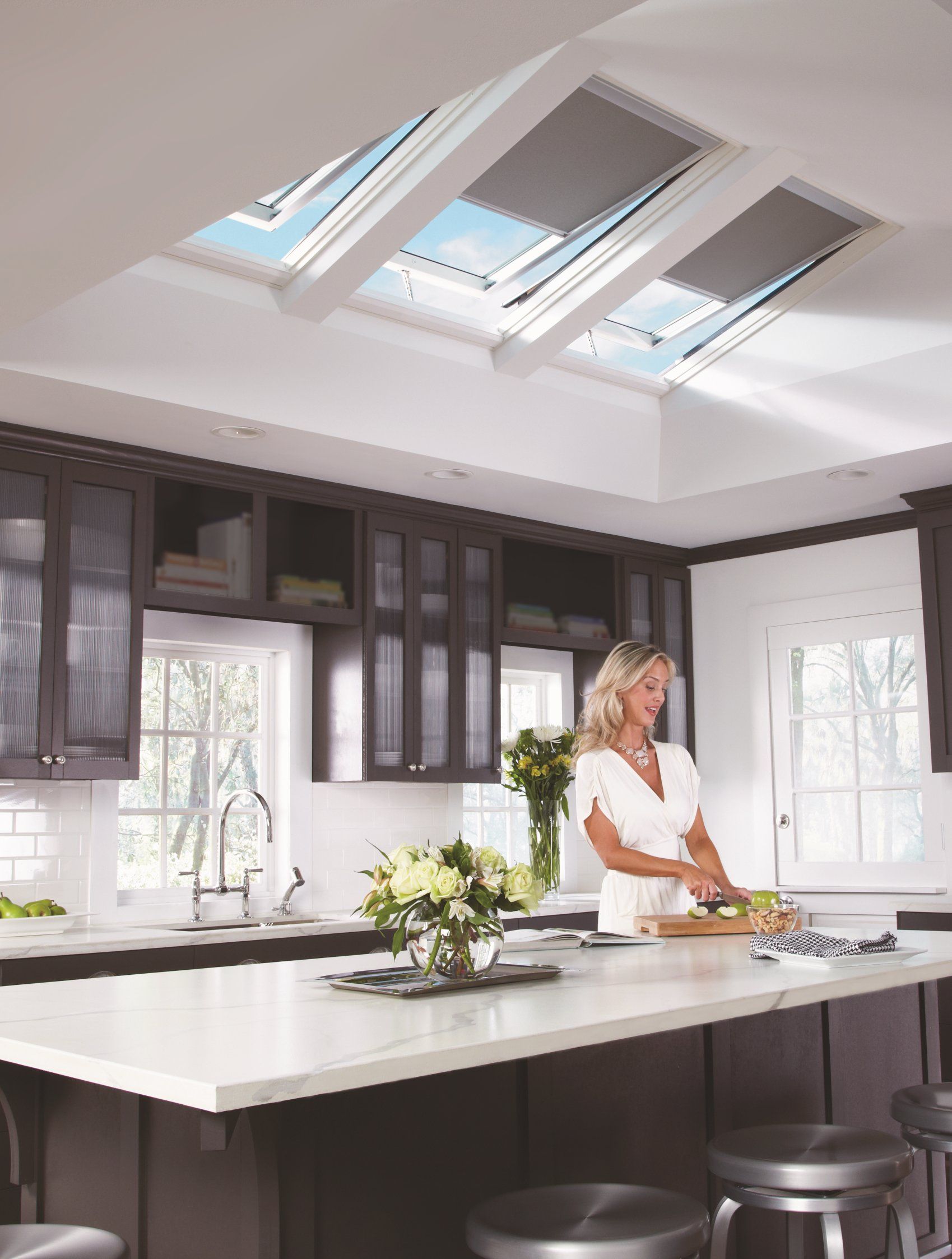 Woman in kitchen with open skylights, chopping food. White countertop, dark cabinets.