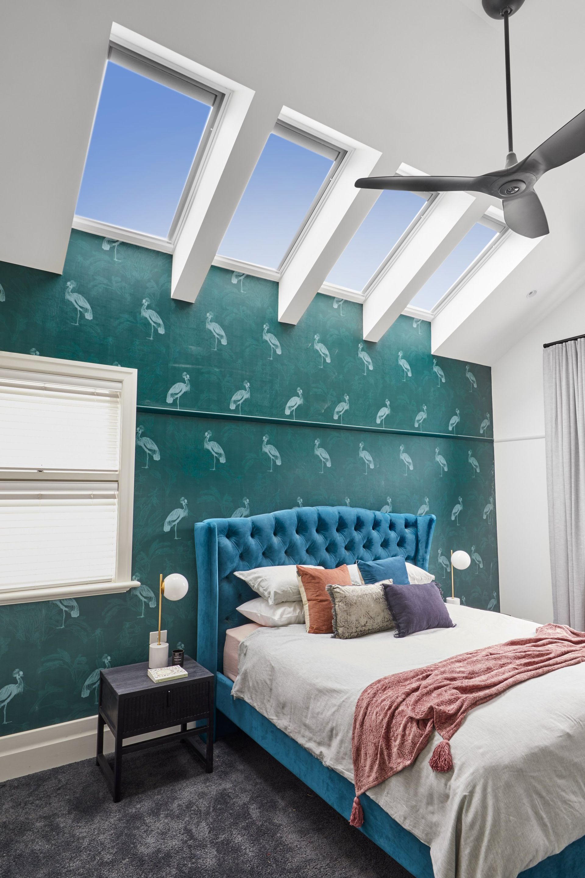 Bedroom with blue velvet bed, teal patterned wallpaper, skylights, and a ceiling fan.