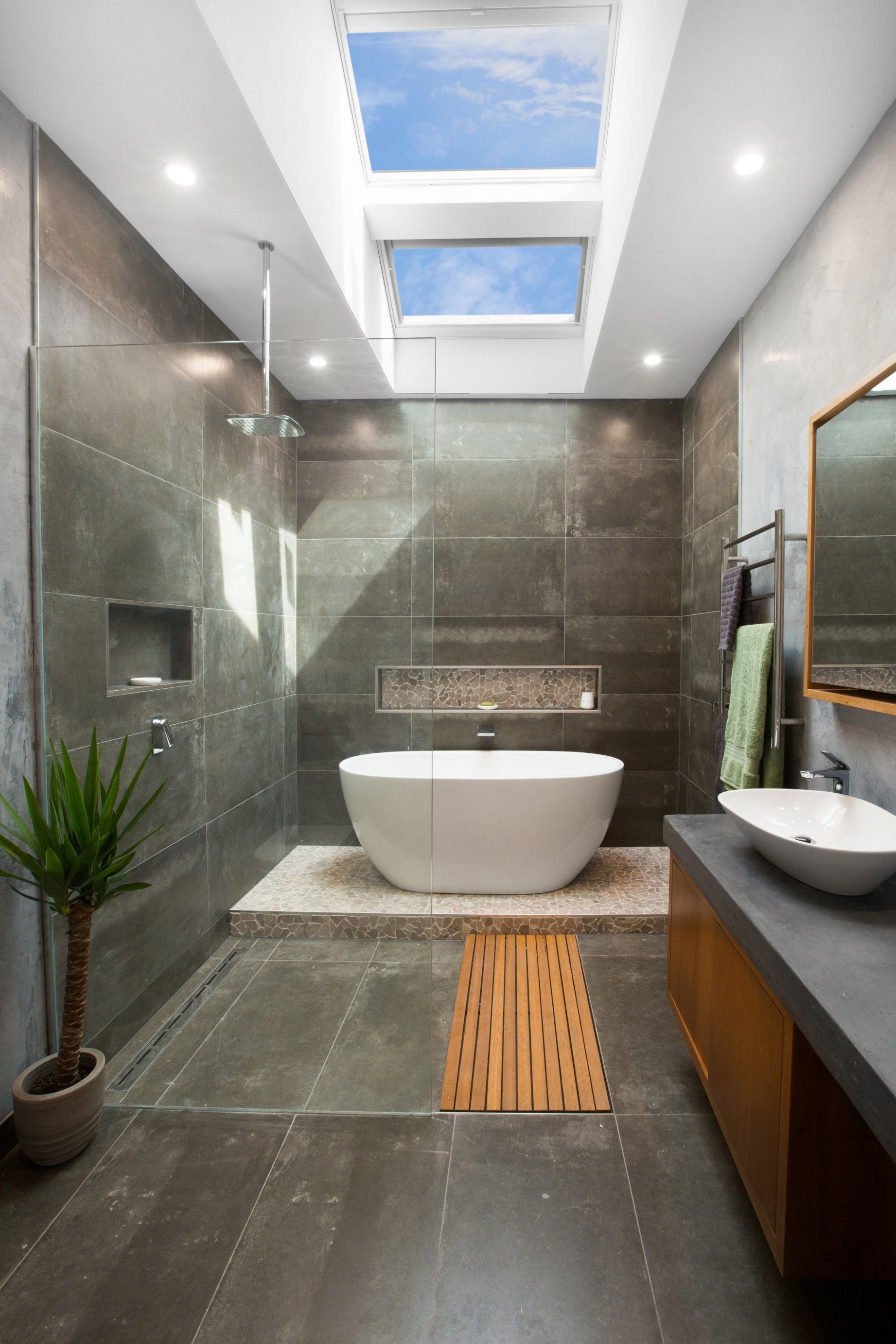 Modern bathroom with skylights, shower, tub, sink, and plant; gray tile and wood accents.