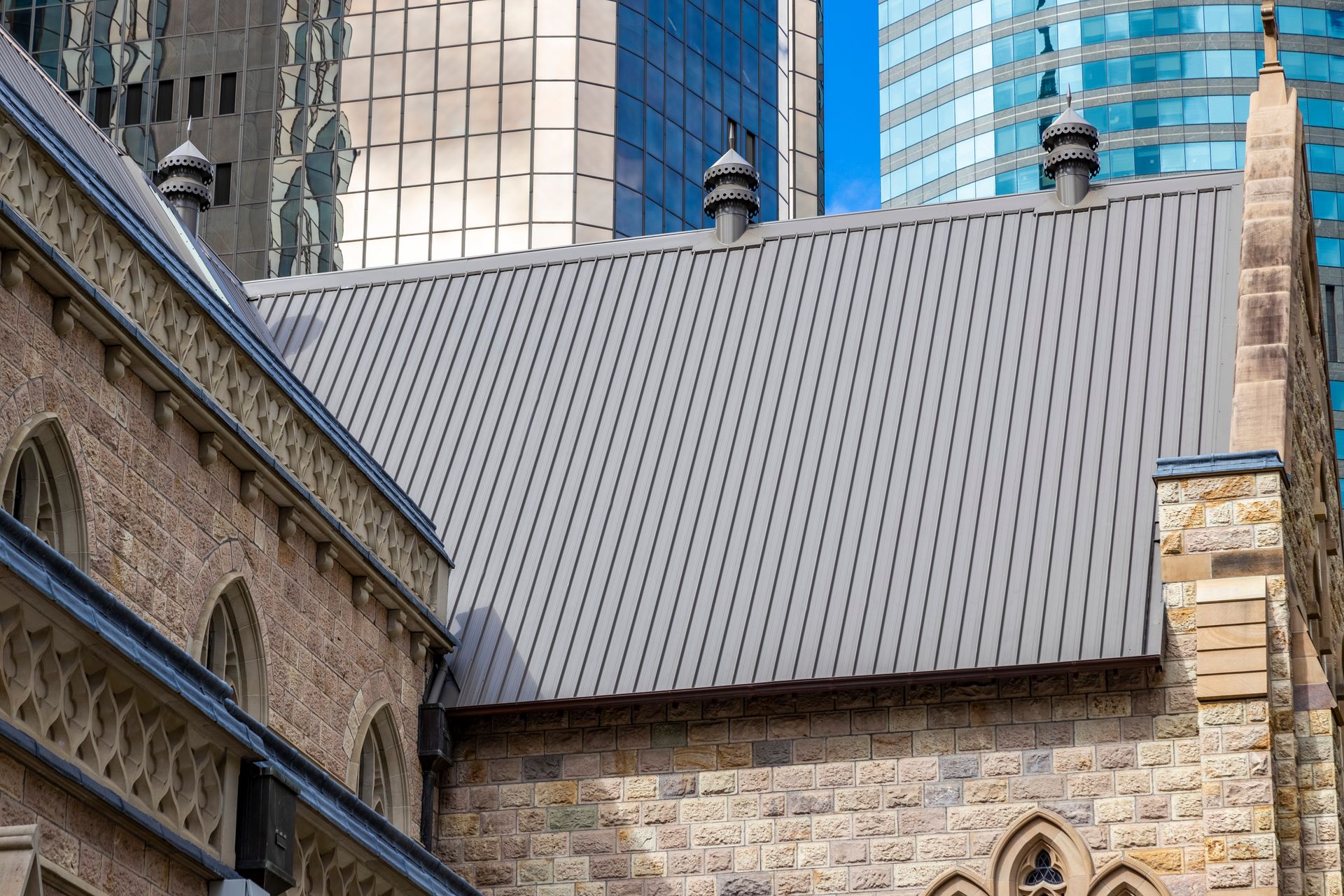 Stone building with a grey metal roof and modern buildings in the background.