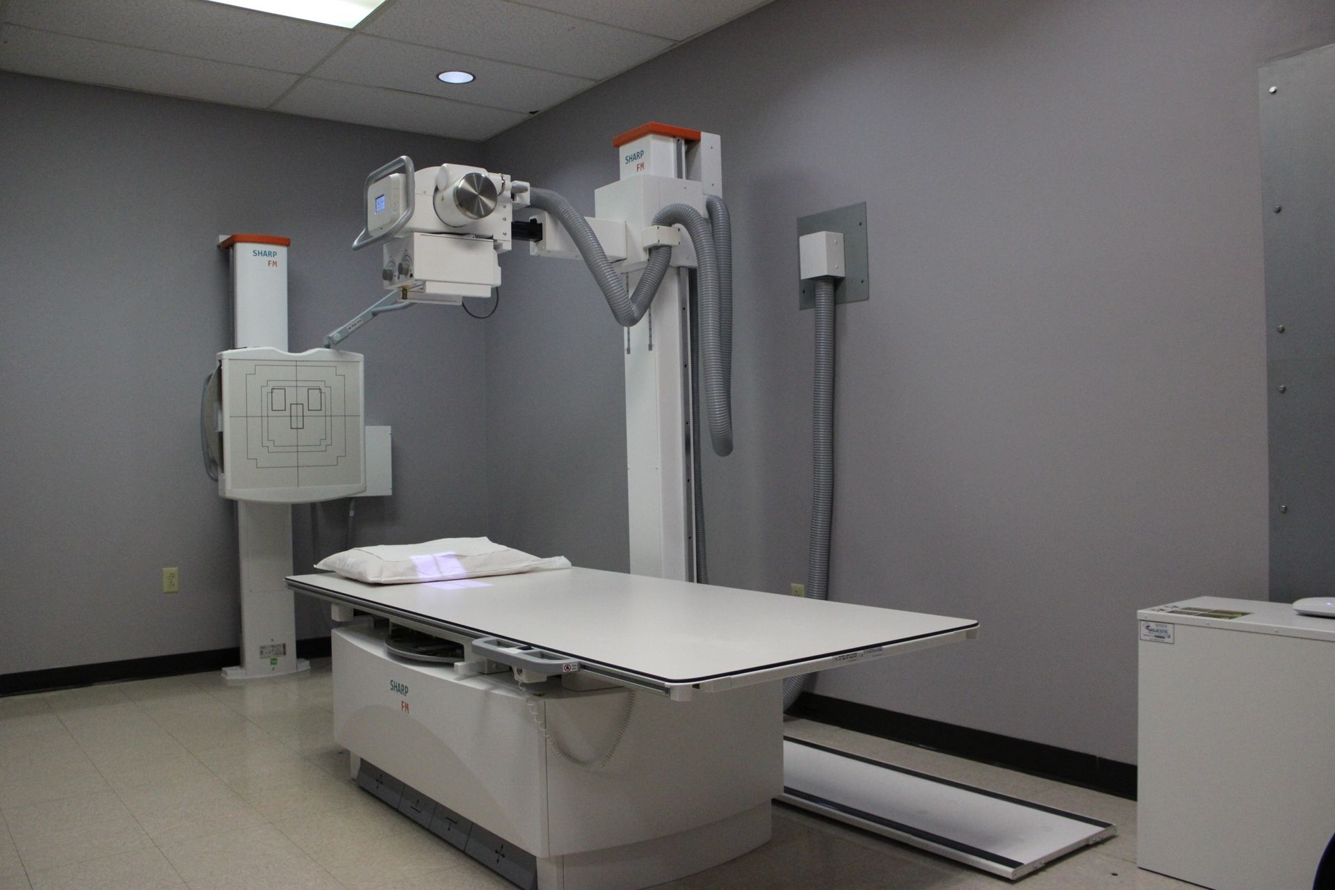An X-ray machine in a gray-walled room. A white examination table is centered beneath the machine.