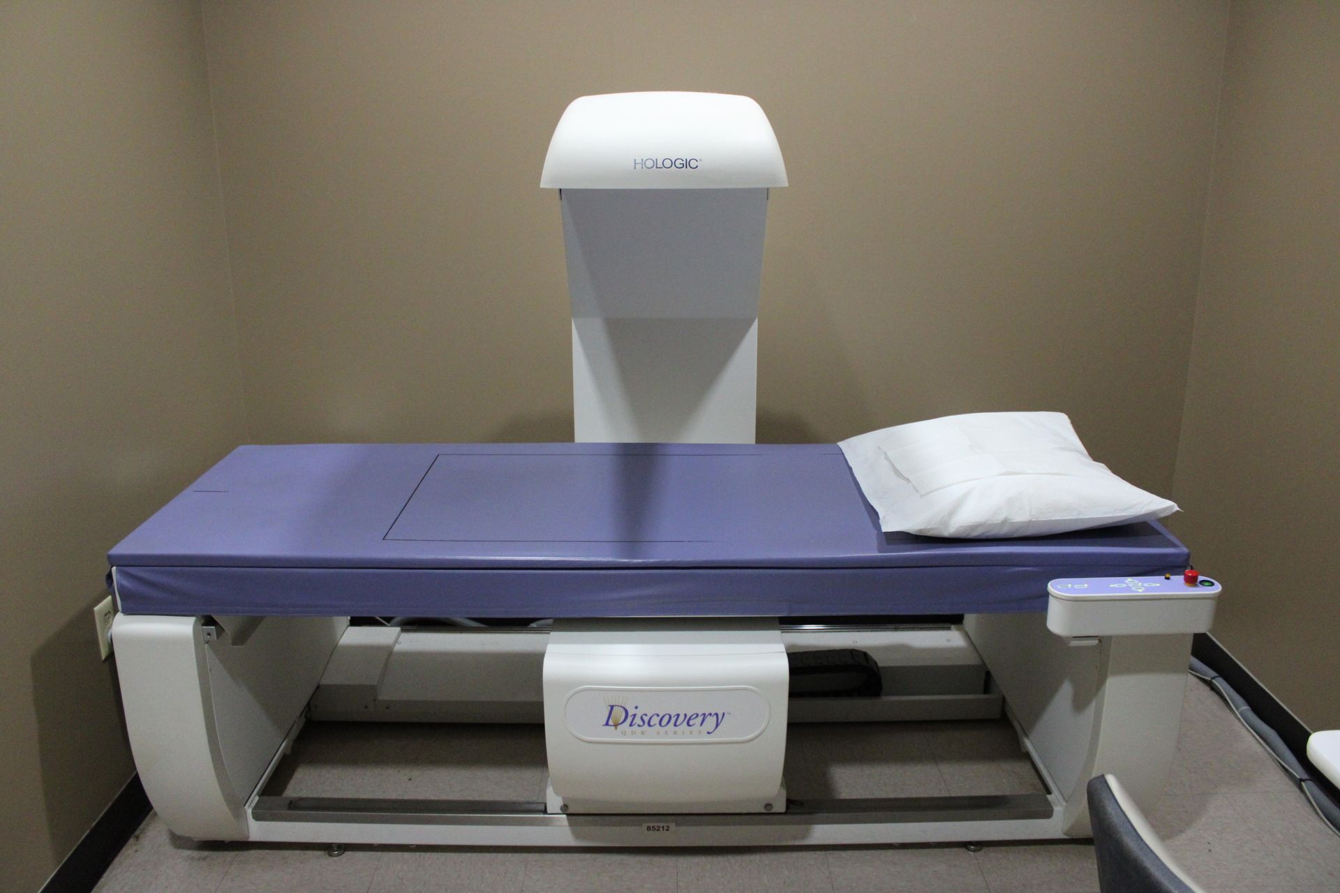 DEXA scanner in a medical examination room, featuring a bed, overhead arm, and pillow.
