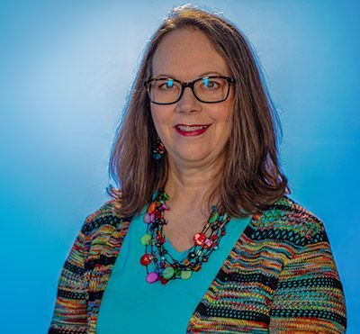 Woman wearing glasses, a colorful necklace, and a patterned sweater smiles against a blue gradient background.