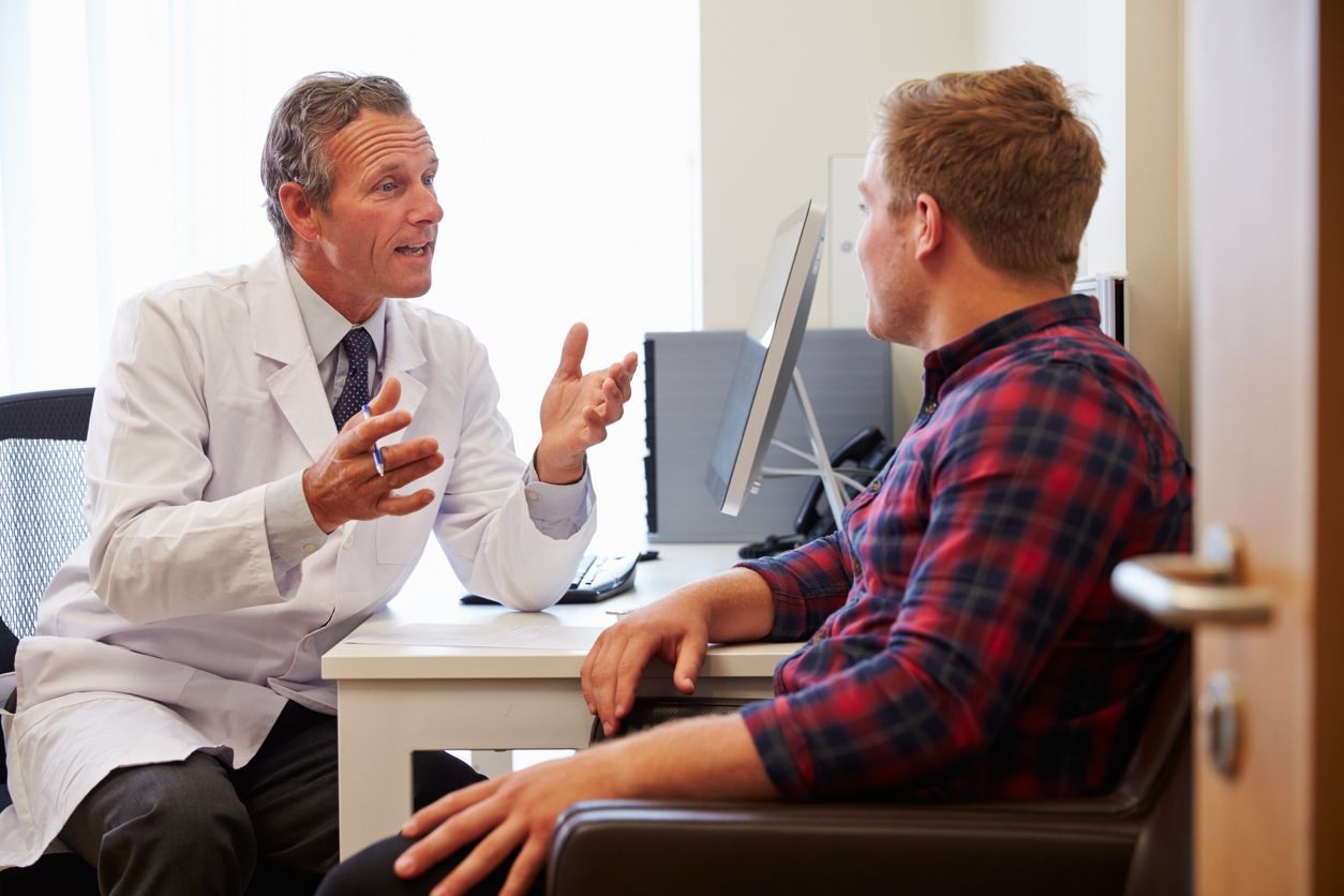 Doctor in lab coat talking to a patient in a plaid shirt at a desk.