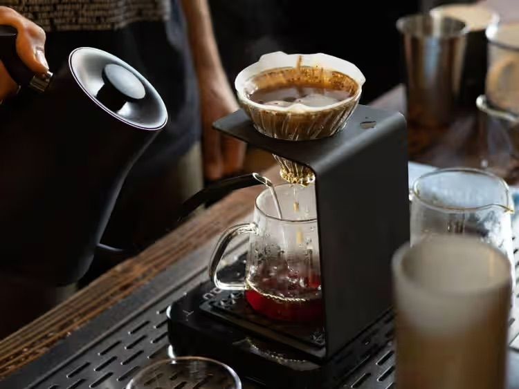 Barista pouring hot water over coffee grounds in a drip brewer, with a glass of milk nearby