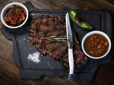 Grilled steak with rosemary, jalapeño, and two side bowls on a black tray with a knife and salt on a wooden table
