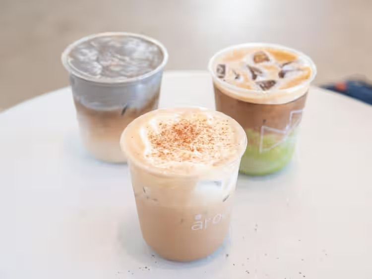 Three iced coffee drinks on a white table, including a tan latte in front and two layered drinks behind.