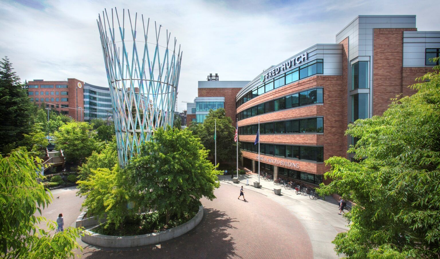 Modern campus courtyard with brick buildings, curved glass facade, and a tall sculpture amid trees