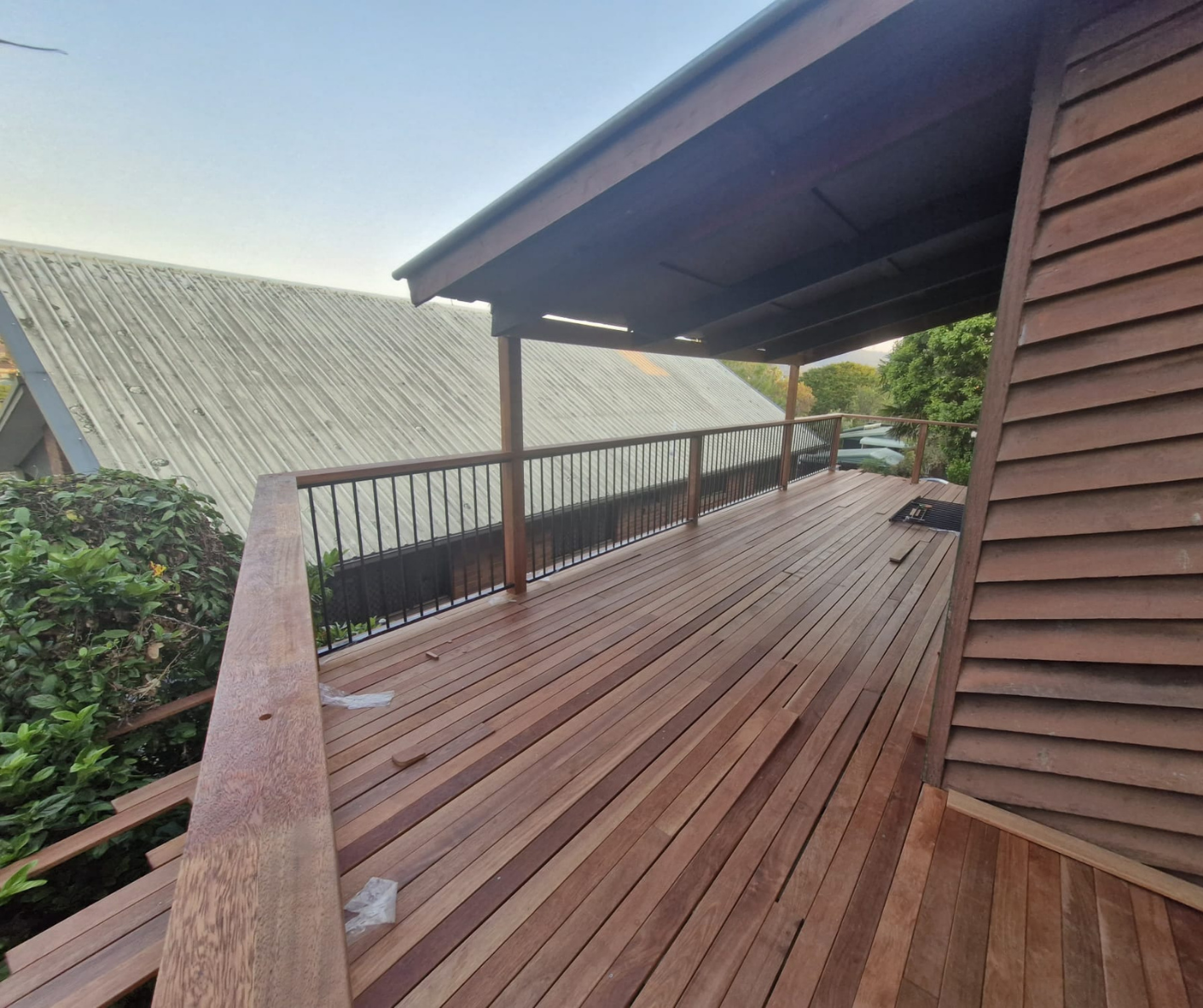 Wooden deck with railing and covered roof beside a house, overlooking greenery and neighboring roofs