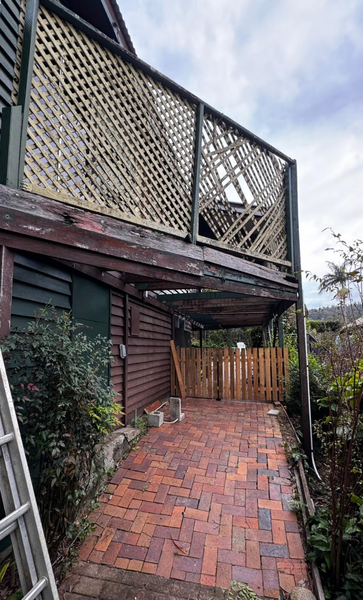 Brick-paved side yard beside a dark wood house with a lattice balcony and fenced gate under a cloudy sky