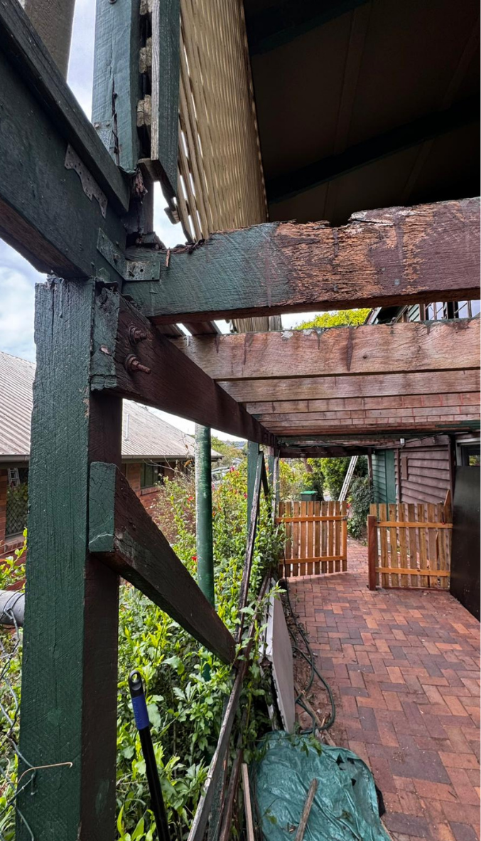 Rusty wooden beams and a brick walkway under a raised porch, with a fence and greenery beyond.
