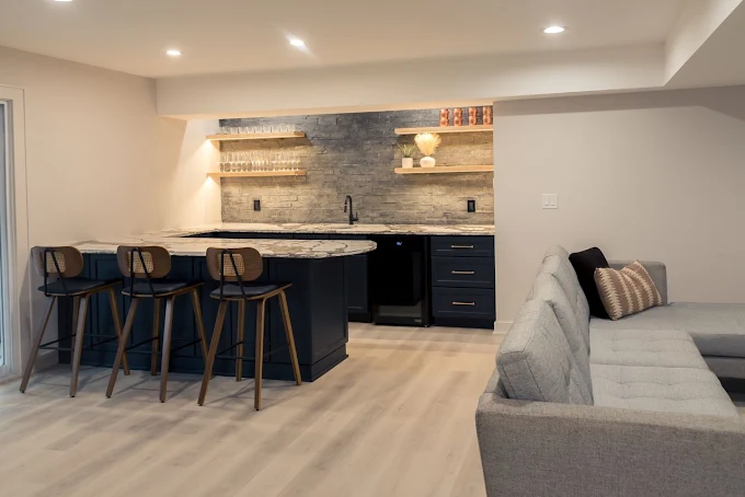 Basement bar with dark blue cabinetry, granite countertop, and wooden stools. Light wood flooring and neutral walls.