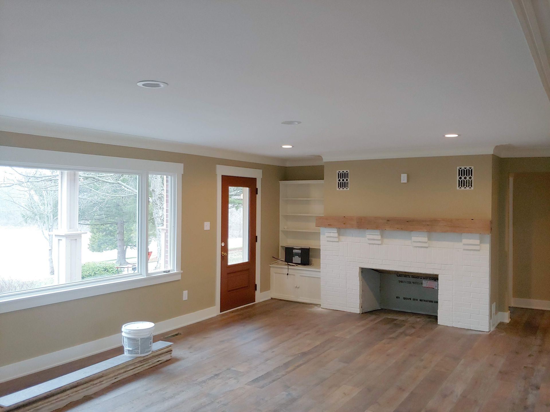A living room featuring white-painted brick fireplace, wood flooring, neutral walls, large window, and a wooden door.
