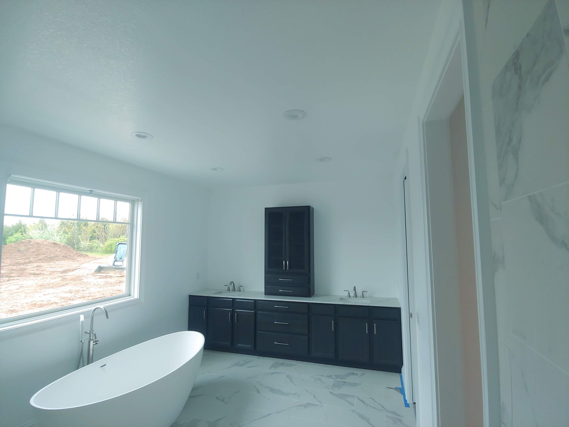A bathroom with a freestanding white tub, dark cabinets, and light marble-patterned tile floors under a bright ceiling.