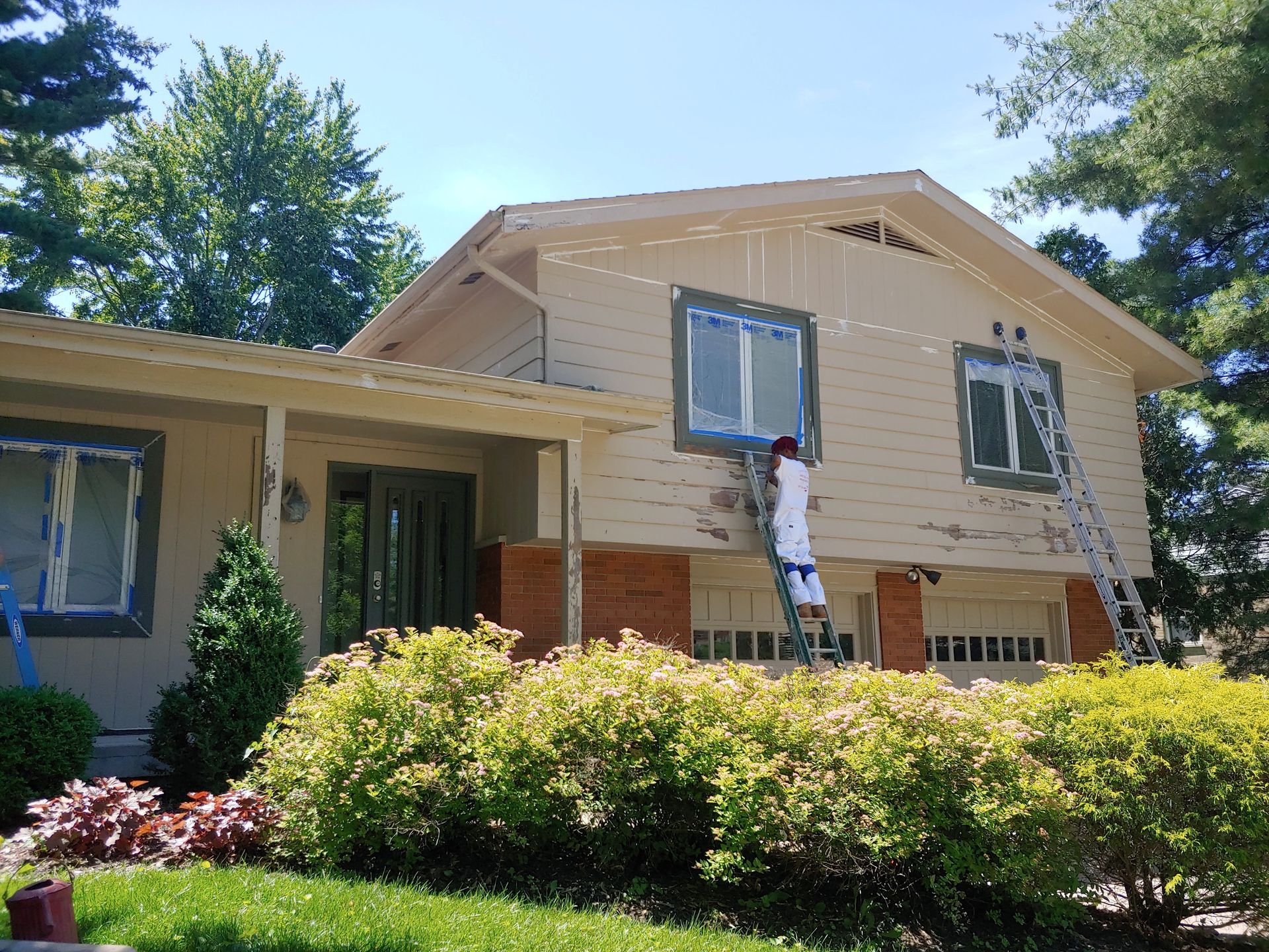 A painter in white clothing on a ladder paints the tan wood siding of a two-story suburban home with brick accents.