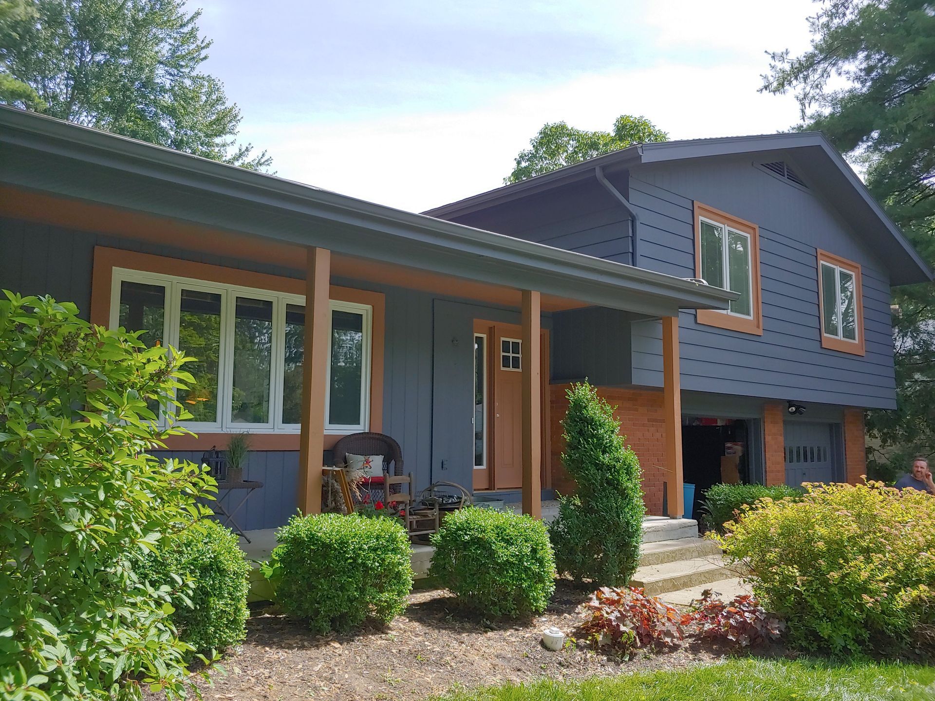 A two-story blue house with wooden trim, a covered front porch, and surrounding landscaping.