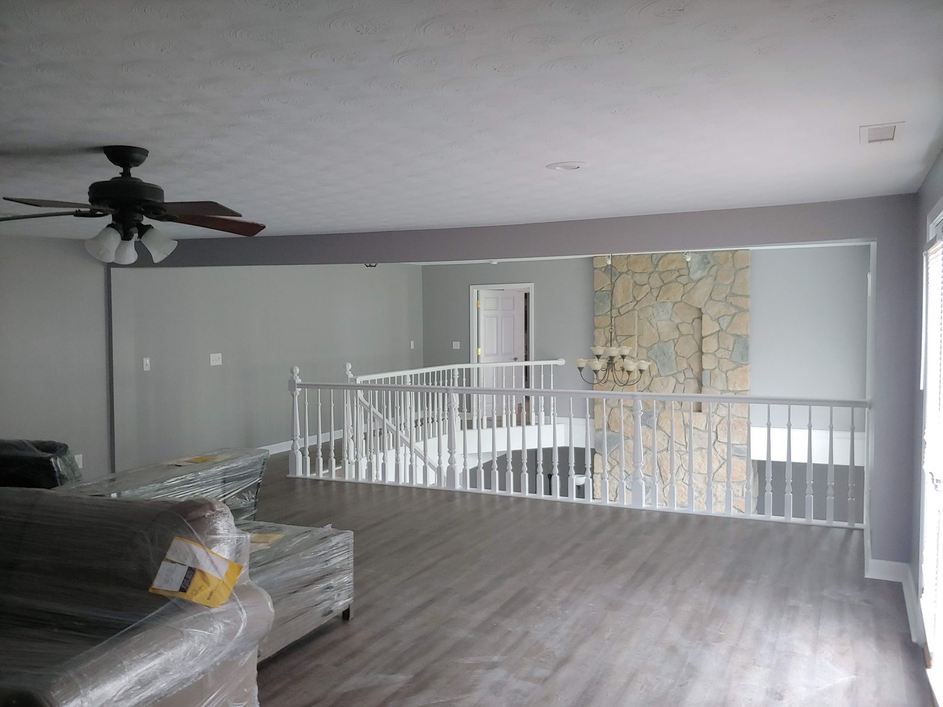 An elevated living room with wood-look floors, a ceiling fan, and a white railing overlooking a foyer with a stone wall.