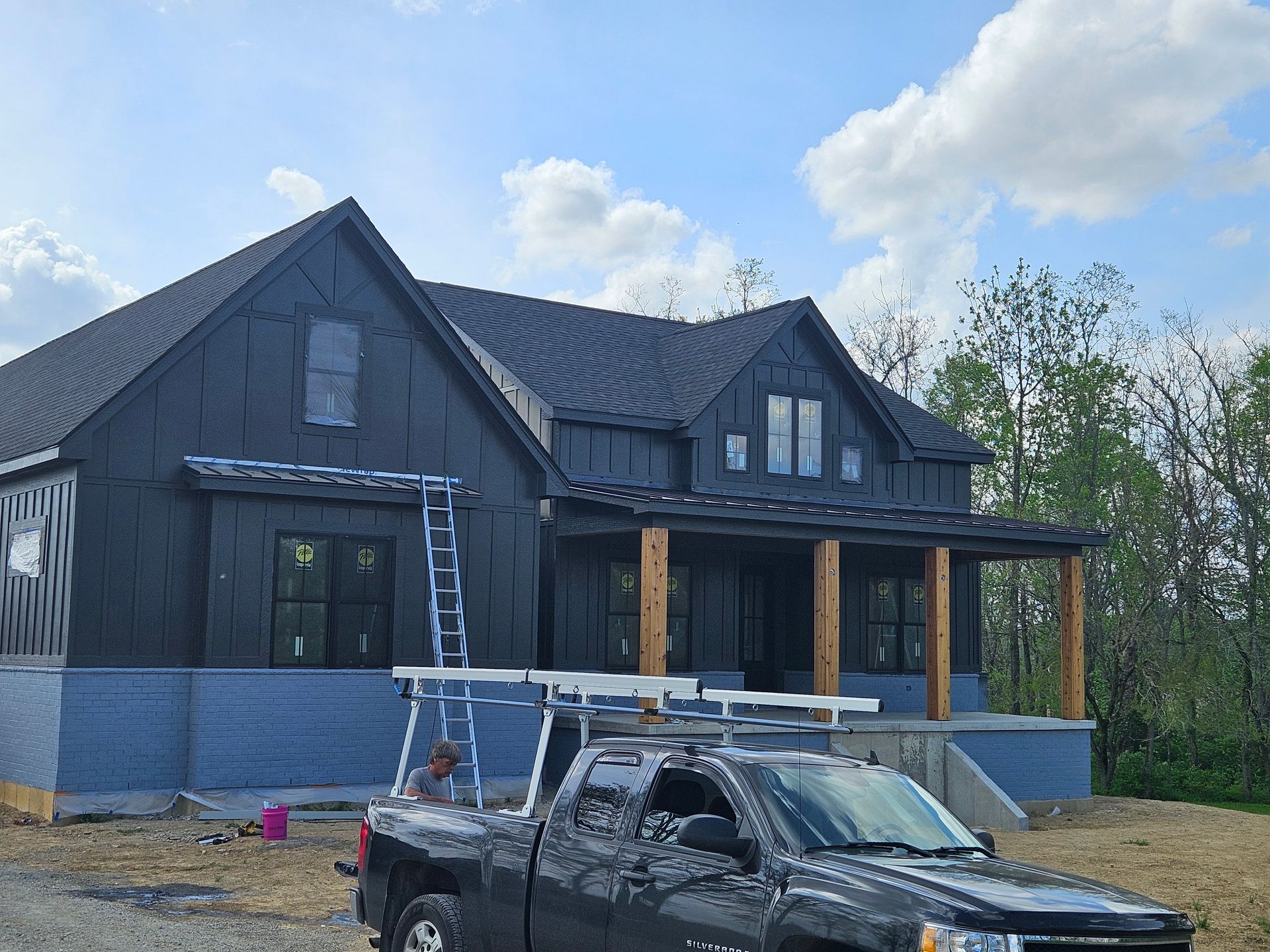 A house under construction with black siding, wood porch posts, and a dark truck parked in front under a blue sky.