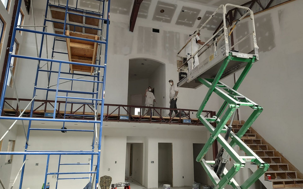 Construction workers in a large, unfinished high-ceilinged room using a blue scaffolding tower and a green scissor lift.