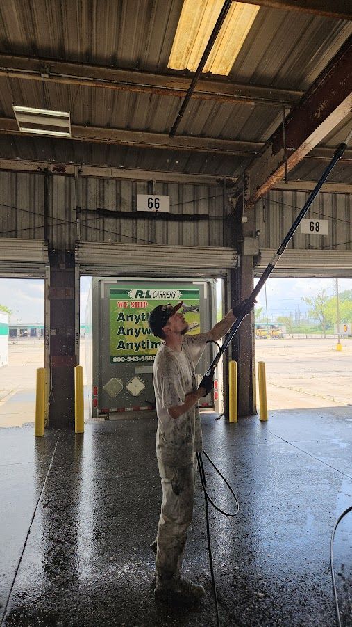 A person in light-colored camouflage clothing uses an extended pressure-washing pole to clean the ceiling of a loading bay.