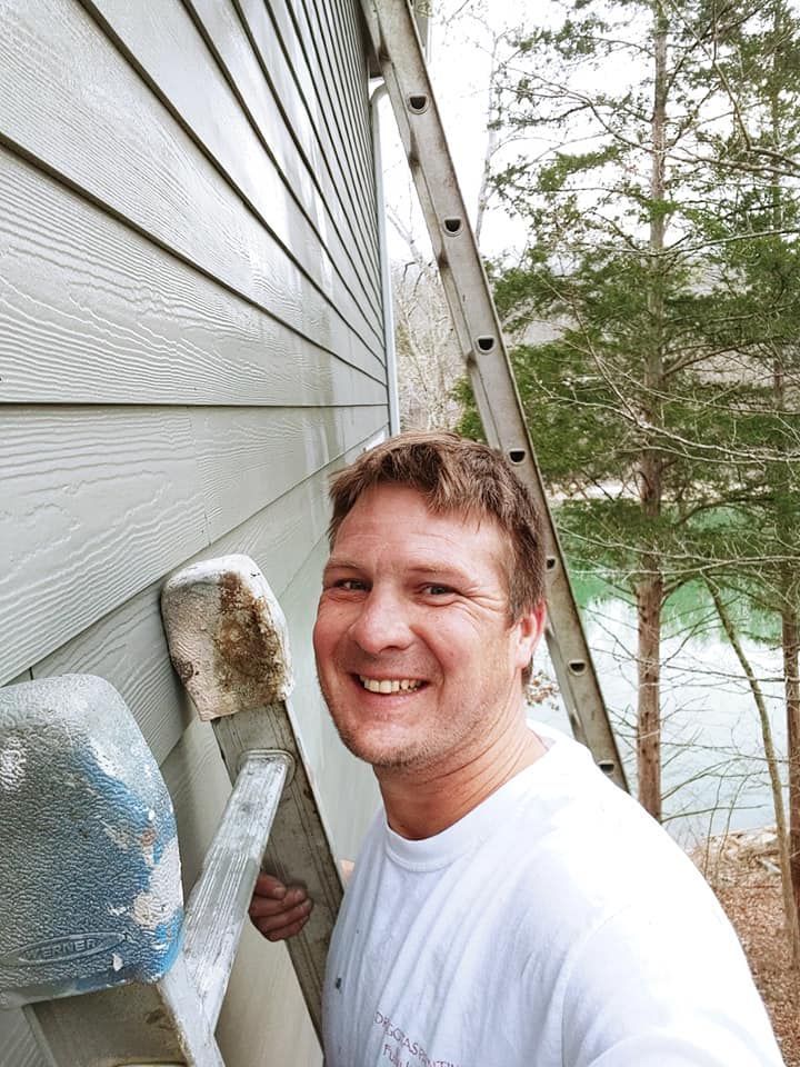 A person smiling while holding a ladder against the side of a house with grey horizontal siding.