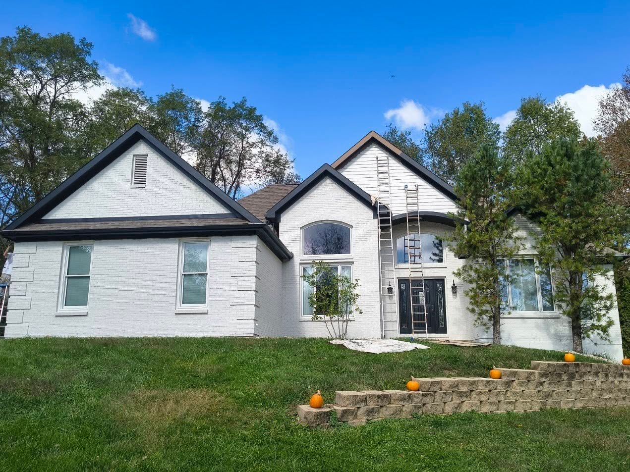 White brick house with a dark roof and black trim, set on a grassy hill with stone steps lined with small orange pumpkins.