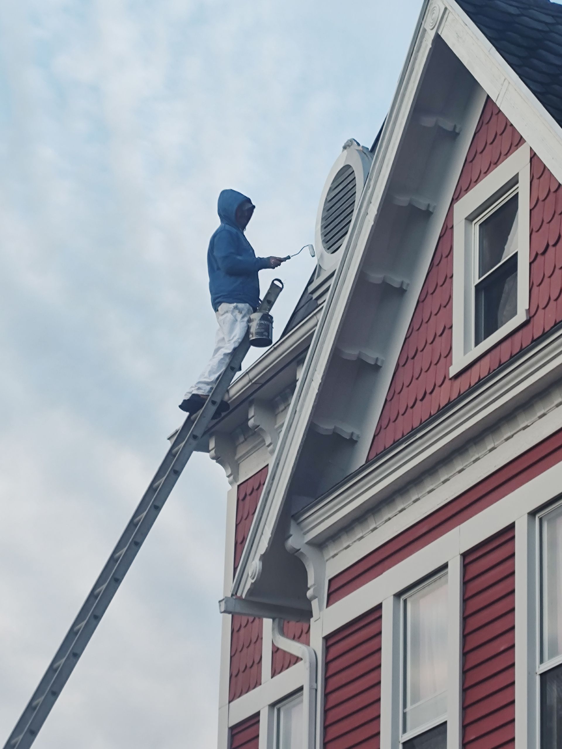 A person in a blue hoodie and white pants stands on a ladder, painting the exterior trim of a red house.