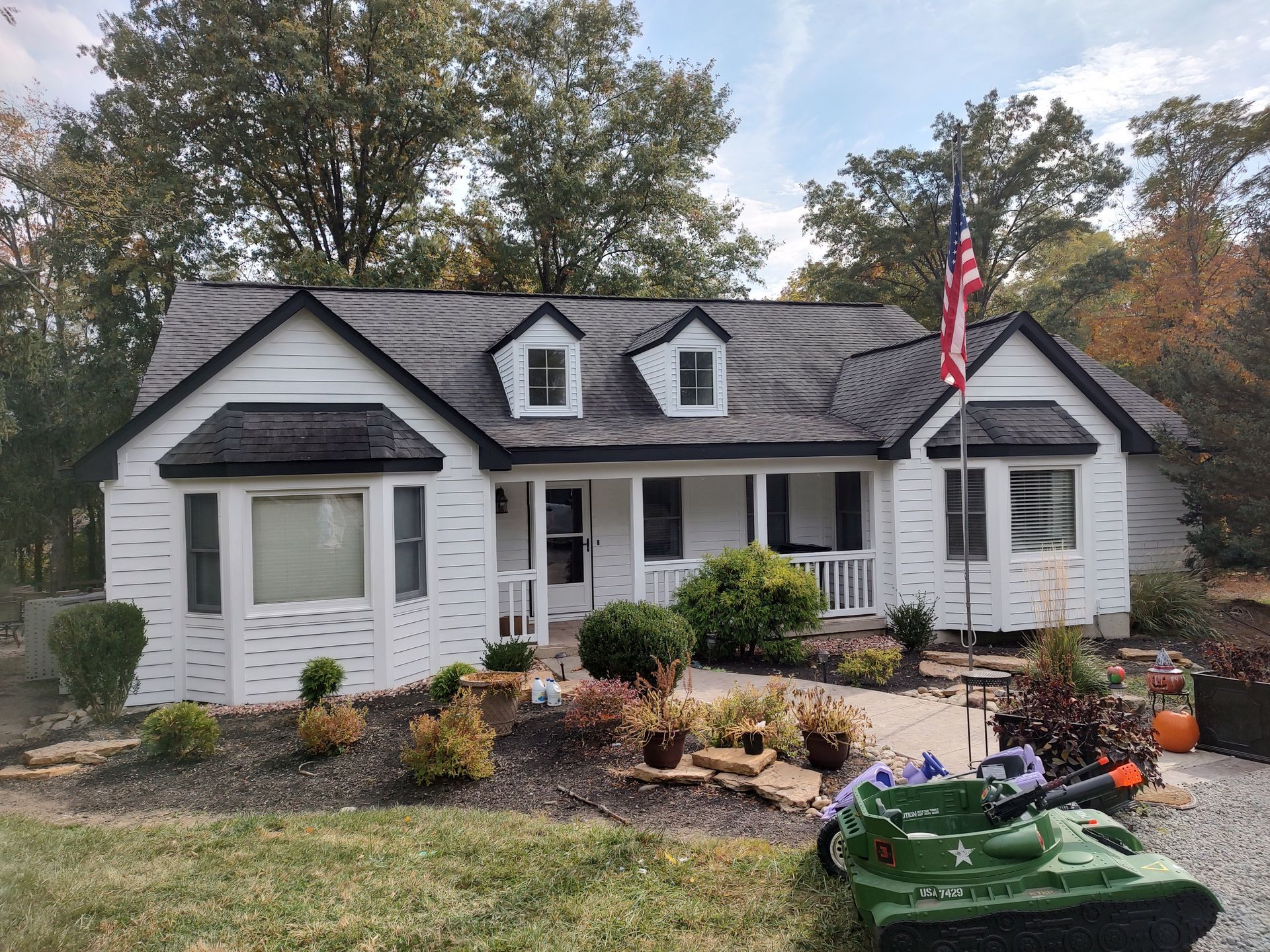 A white, one-story house with dark trim, dormer windows, and a green toy tank in the front yard under an American flag.