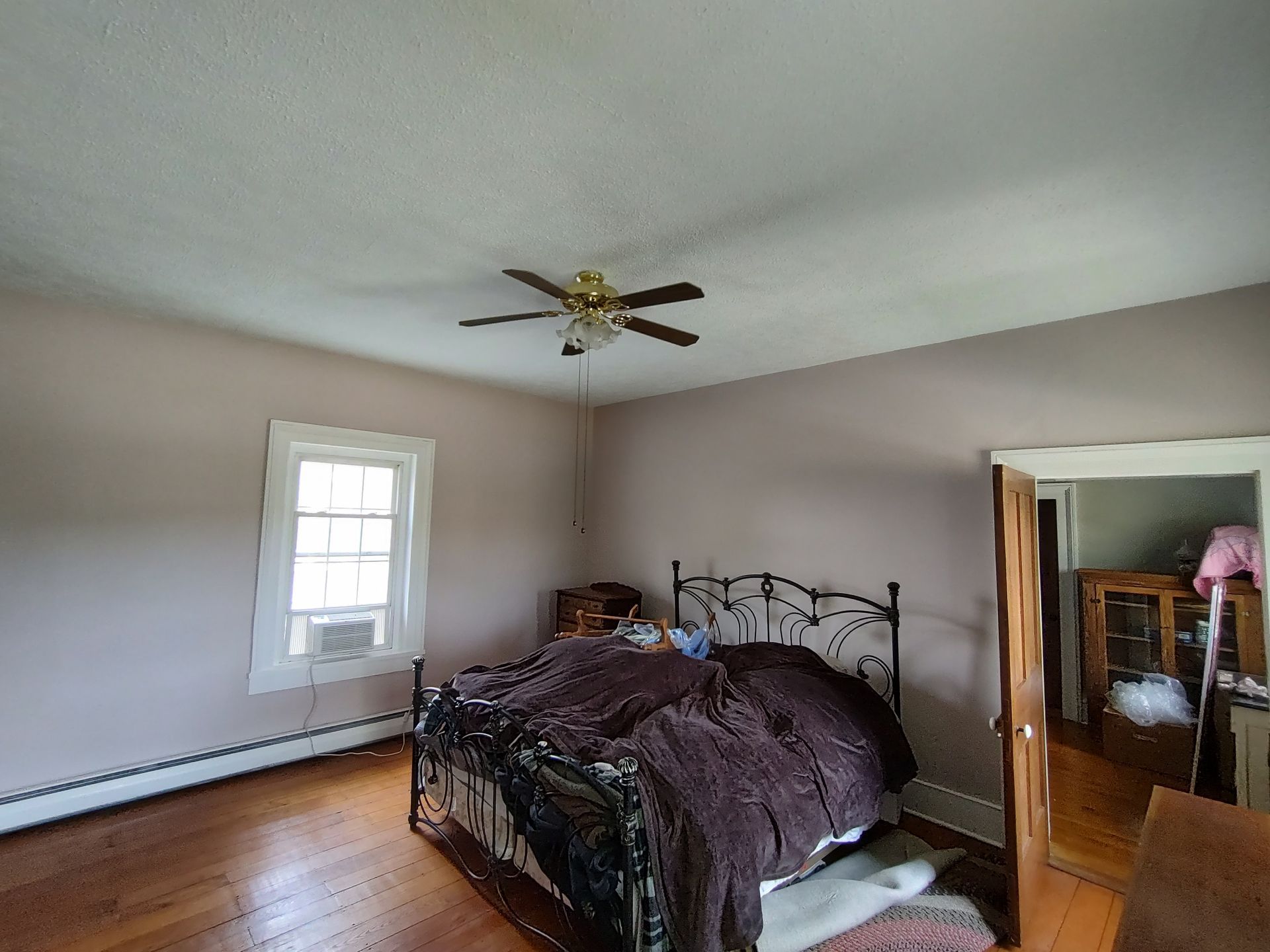 A bedroom with wooden floors, pale walls, a metal bed frame with a dark bedding, a ceiling fan, and a window.