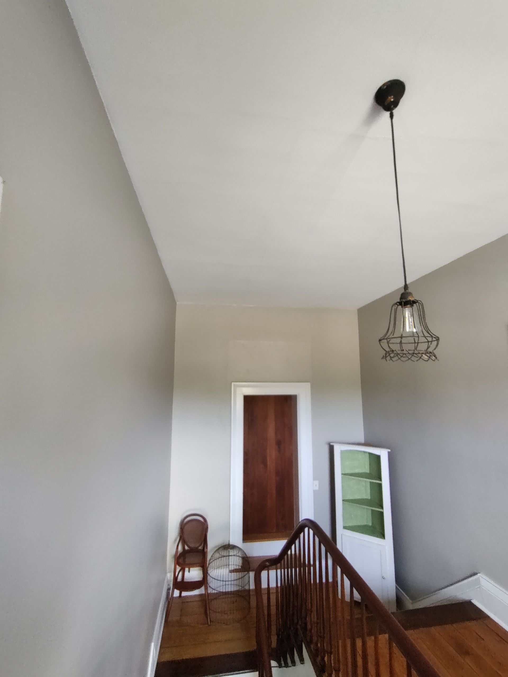 A view from a wooden staircase looking up at a hallway with light gray walls, a pendant light, a small chair, and a cabinet.