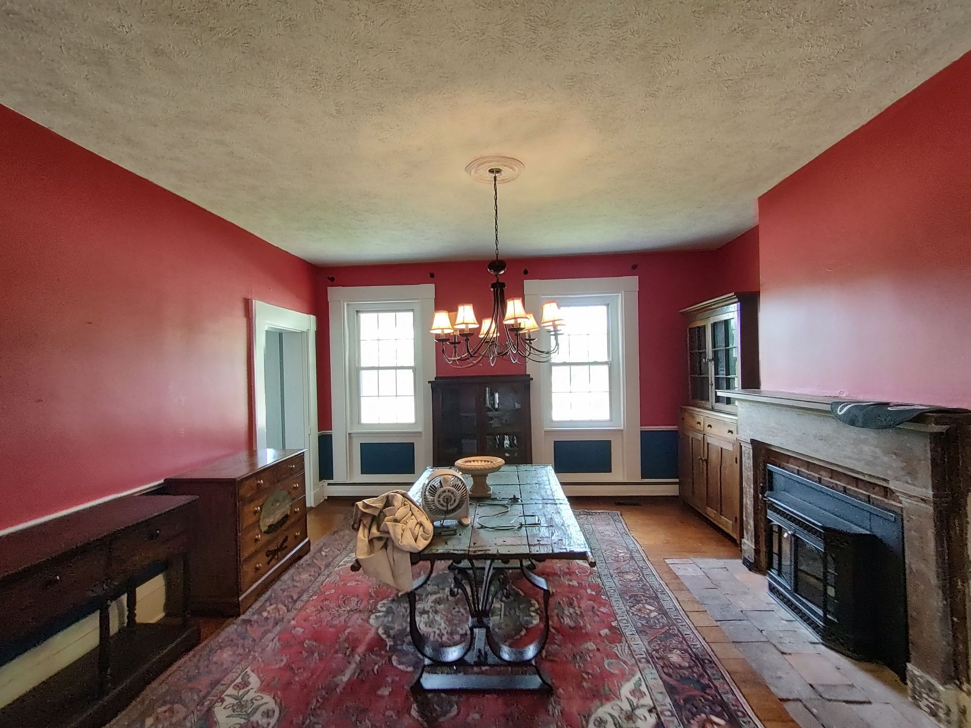 A dining room with red walls, a large wooden table, a fireplace, and a patterned area rug under a chandelier.