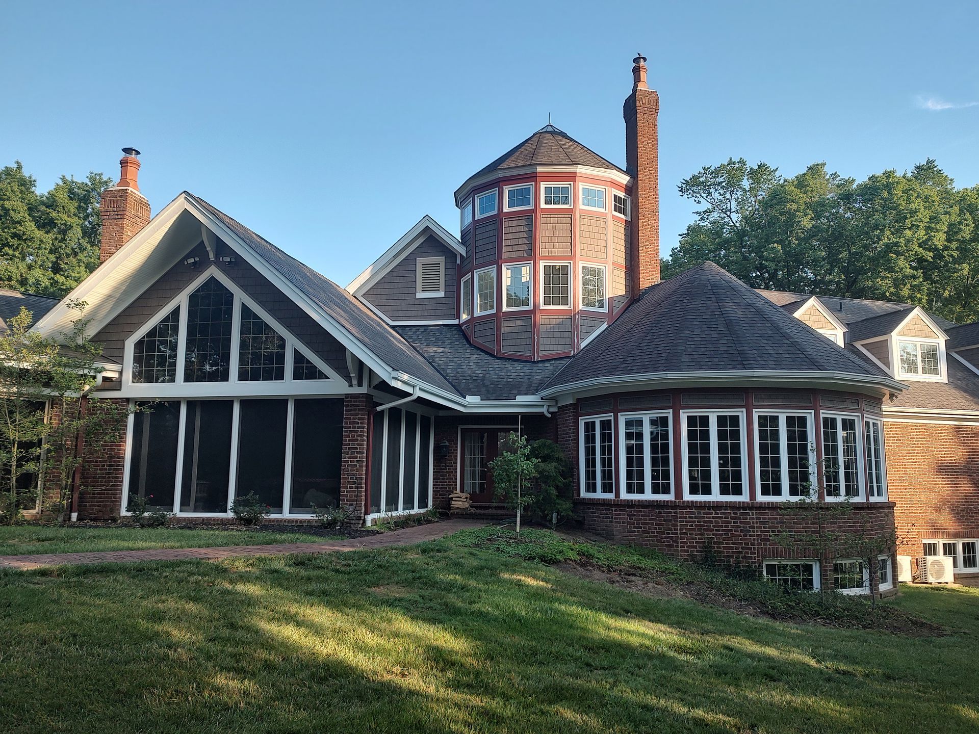 A large, multi-story house with brown shingle siding, a central turret, large windows, and a grassy yard under a blue sky.