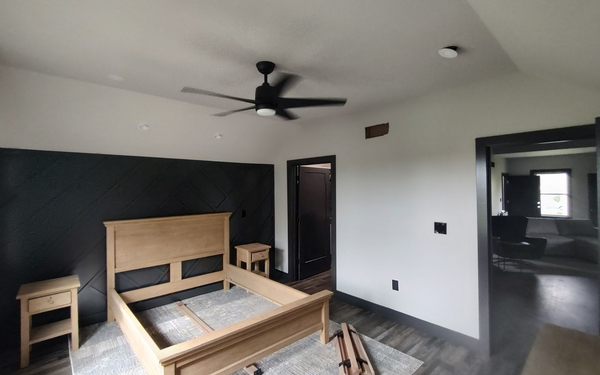 A modern bedroom with light wood bed frame, black accent wall, ceiling fan, and two doorways leading to other rooms.